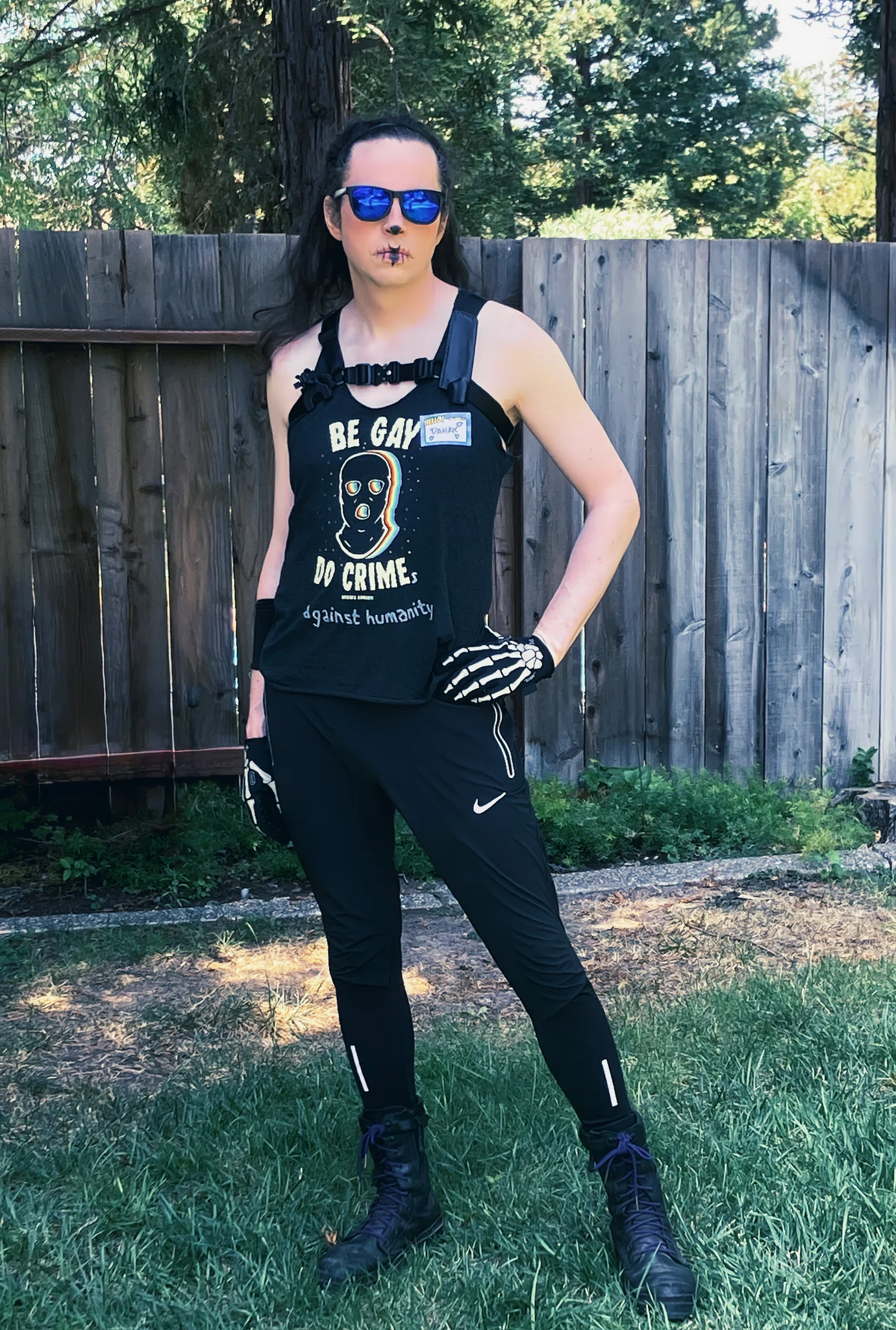 The cavalier primary of the Ninth House to serve the King Undying stands before you in a power pose, shoulders back and hips cocked, skeleton-gloved-hands resting easy but ready at their sides. Long hair trails majestically over their bare shoulders above a "Be Gay Do Crime" tank top with "against humanity" doodled on as if an afterthought. A chest and shoulder harness supports holsters & sheathes for several weapons which seem to be missing. Their face has only a hint of black around the lips and nose, parsimonious suggesting the visage of a skeleton. Even behind reflective blue lenses, the viewer can feel this cavalier's eyes sizing them up & assessing them: friend or foe; threat or… opportunity?